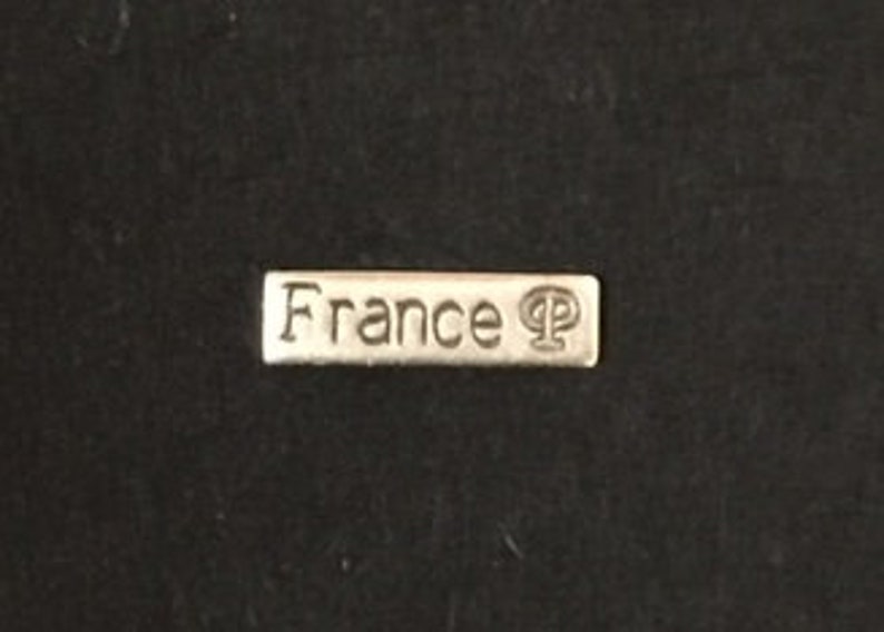 以下が含まれることがあります： クラシックなフォントで「France」の文字が刻まれた、小さく長方形のシルバー調の金属タグ。タグには小さなシンボルも付いています。タグは暗い、おそらく黒の背景に配置されています。