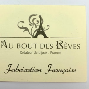 以下が含まれることがあります： クリーム色の長方形のタグで、黒い文字とエッフェル塔のグラフィックが描かれています。「Au Bout des R&ecirc;ves, Cr&eacute;ateur de bijoux, France」と「Fabrication Fran&ccedil;aise」と書かれています。
