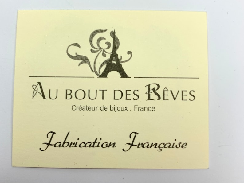 以下が含まれることがあります： クリーム色の長方形のタグで、黒い文字とエッフェル塔のグラフィックが描かれています。「Au Bout des R&ecirc;ves, Cr&eacute;ateur de bijoux, France」と「Fabrication Fran&ccedil;aise」と書かれています。