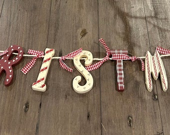 May include: A wooden Christmas banner with letters in alternating colors of cream and red. The letters are strung together with a white cord and red and white checkered ribbon. The banner is displayed on a wooden surface.