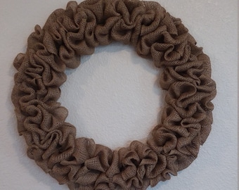 May include: A brown burlap wreath with a ruffled design. The wreath is circular and hangs against a white wall. The burlap material has a natural, textured appearance, creating a rustic aesthetic.