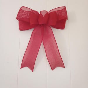 May include: A large, vibrant red bow made of sheer, textured ribbon. The bow features multiple loops and two long, pointed tails. The bow is likely used for decoration, such as on a gift or wreath. The background is a plain white surface.
