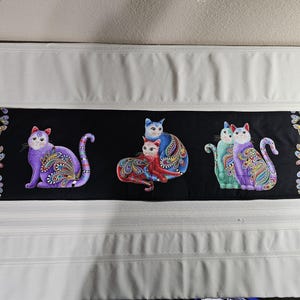 May include: A black fabric runner with colourful cat illustrations. The cats are in various poses and adorned with intricate paisley patterns. The runner has decorative borders with floral and paisley designs.