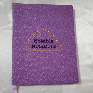 以下が含まれることがあります： 紫色のジャーナル。ドット模様があり、「Notable Notations」の文字が黒と金で刺繍されています。ジャーナルにはリボンブックマークと、テキストの上に金の星のデザインがあります。