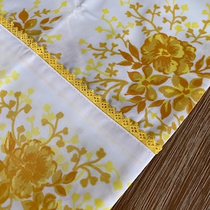 May include: A white fabric with a yellow floral pattern and a yellow crocheted trim. The fabric is folded in half and the trim is visible on the edge.