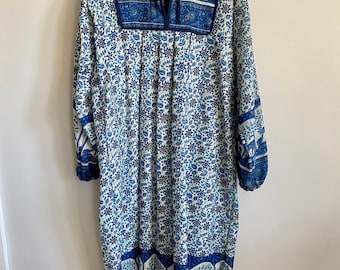 May include: A long-sleeved blue and white floral print dress with a tie-up neckline and a blue embroidered yoke. The dress has a gathered skirt and a blue and white border with a repeating elephant design.