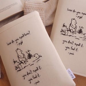 May include: Two cream-coloured book covers featuring a Winnie the Pooh illustration and quote. The quote reads, "How do you spell love? - Piglet. You don't spell it, you feel it. - Pooh." An open book and a sweater are also in the image.