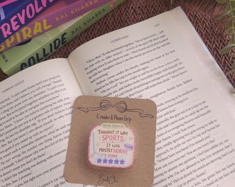 May include: A square e-reader and phone grip on a brown cardboard backing, featuring a pink and white graphic with the text "BOOK REVIEW", "THOUGHT IT WAS SPORTS", "IT WAS MOSTLY HORNY", and "5 STARS" with five purple stars and a hockey stick icon.