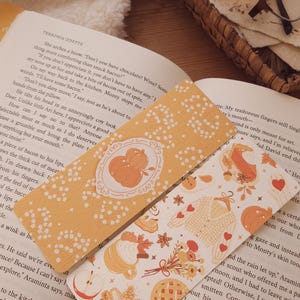 Puede incluir: Dos marcapáginas decorativos con diseños otoñales. Uno presenta una ilustración de calabaza dentro de un marco decorativo, el otro muestra hojas, corazones y alimentos como gofres y pastel. Los marcapáginas están sobre un libro abierto.
