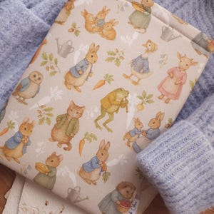 Può includere: Una copertina di libro beige con illustrazioni di Peter Coniglio e altri personaggi. La copertina presenta un motivo ripetuto di conigli, anatre e altri animali, con carote e annaffiatoi. La copertina è appoggiata su un maglione blu.