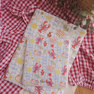 Può includere: Una custodia protettiva trasparente per tablet con un motivo ripetuto di aragoste rosse, fette di limone giallo, erbe verdi e scatolette di sardine su sfondo a griglia azzurro, con il testo "la dolce vita" e "Book Chic" stampato sopra.