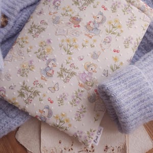 May include: A book cover with a whimsical pattern of ducklings in bonnets, butterflies, and flowers. The cover is cream-colored with a dotted texture. A light blue sweater is in the background. The cover has the text "Book Chic".