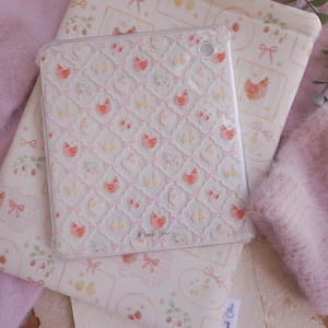 Puede incluir: Una funda transparente para tableta con un patrón repetitivo de pollos pequeños, pollitos y lazos rosas dispuestos en una cuadrícula. La funda tiene el texto BookChic impreso en cursiva en la parte inferior central y un recorte circular.