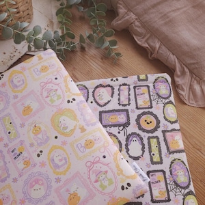 Peut inclure: Deux pièces de tissu avec un motif d'Halloween avec des fantômes, des citrouilles et des cadres aux couleurs pastel. Le tissu est rose clair et violet. Un coussin beige avec un bord à volants et un panier en osier avec de la verdure sont en arrière-plan.