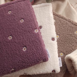 May include: Three plush, textured book sleeves in purple, cream, and brown, each embroidered with small white daisies. The sleeves are stacked, with a book visible in the background.