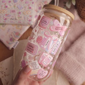 May include: A clear glass tumbler with a wooden lid and a glass straw. The tumbler is decorated with pink stickers featuring the words "Hockey Romance," hearts, bows, and other romantic elements. The tumbler is held in a hand.