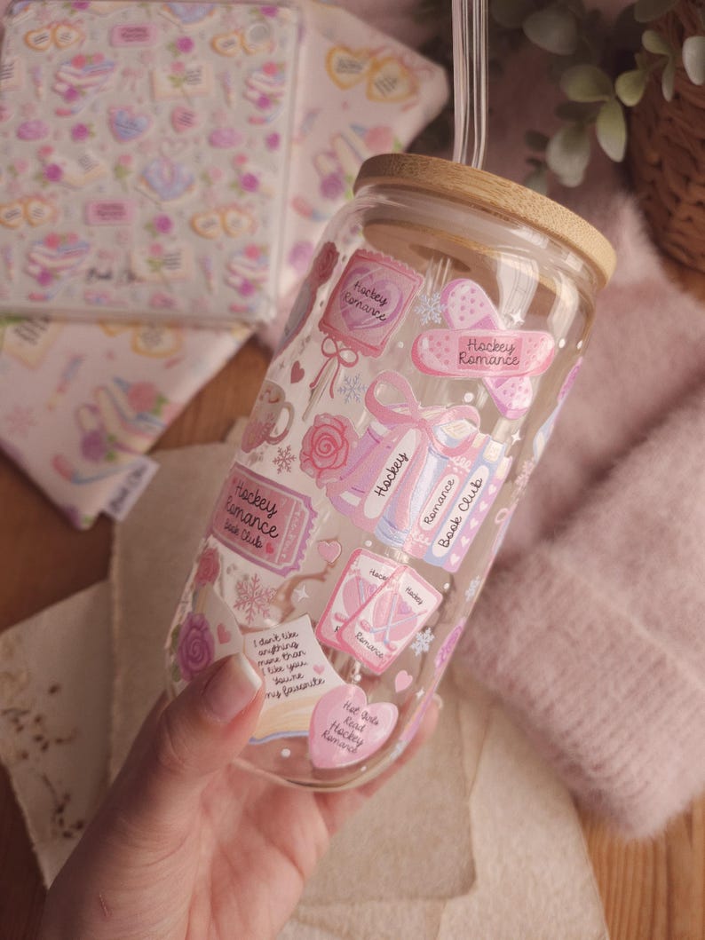 May include: A clear glass tumbler with a wooden lid and a glass straw. The tumbler is decorated with pink stickers featuring the words "Hockey Romance," hearts, bows, and other romantic elements. The tumbler is held in a hand.