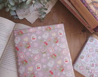 Puede incluir: Una funda de libro lila con un patrón repetido de cerezas, tazas de té, libros y flores. La funda está junto a un libro abierto y una pila de libros en una cesta de mimbre. Una pequeña planta está en el fondo.