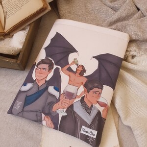 以下が含まれることがあります： 3 人の人物が描かれたカラフルなイラストが施された白い本のカバー。人物はファンタジーな服装をしていて、翼があります。カバーには「Book Chic」と書かれたラベルが付いています。