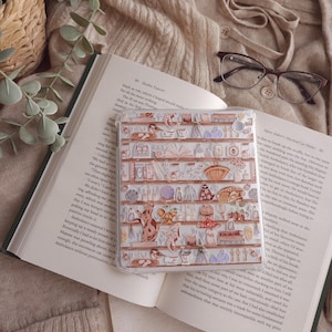 May include: A transparent phone case with a detailed illustration of shelves displaying cats, teacups, and other decorative items. The case is placed on an open book, with a beige sweater and spectacles in the background.
