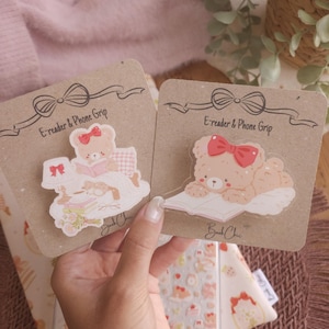 May include: Two acrylic e-reader and phone grips on brown cardboard backing cards. One grip features a bear reading in bed with a lamp and books. The other grip shows a bear lying down reading a book. Both bears wear red bows. Text reads BookChic.