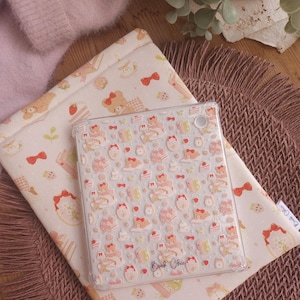 Puede incluir: Una funda transparente para tableta con un patrón repetitivo de ositos, pasteles y lazos rojos, con el texto Book Chic impreso en la parte inferior. La funda descansa sobre una funda de tela con un estampado a juego de osos, lazos y fresas.