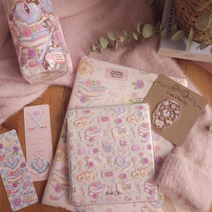 May include: A collection of pink and white stationery and accessories, including a tumbler, tablet case, stickers, and a phone grip. The items feature a repeating pattern of books, roses, and ice skates. The text "Hockey Romance Book Club" is visible.