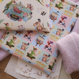 Op de afbeelding: Verschillende stoffen items met Peter Pan-personages en scènes. Een etui toont een vintage illustratie van Peter Pan's Neverland Adventure. Een ander item heeft een herhalend patroon van Peter Pan en andere personages. De items zijn in crème, roze en blauwe tinten.
