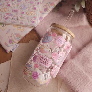 May include: A clear glass tumbler with a bamboo lid and a pink silicone sleeve. The tumbler is decorated with stickers featuring hearts, roses, and the text "Hockey Romance Book Club". A clear straw is inserted into the lid. A matching book cover is in the background.