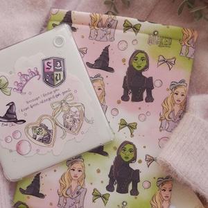 Puede incluir: Una funda blanca para tableta y una funda de tela con ilustraciones del musical "Wicked". La funda tiene una cita y un escudo escolar. La funda tiene imágenes de la Bruja Malvada y Glinda, con la palabra "Wicked" impresa.