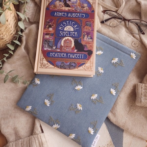 May include: A book titled "Agnes Aubert's Mystical Cat Shelter" with a blue book sleeve featuring white daisy embroidery. The book and sleeve rest on a beige sweater. The book cover has illustrations of cats.