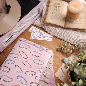 Può includere: Tre copertine di libri rosa con un motivo di braccialetti di perline colorate e i nomi degli album di Taylor Swift: "Reputation", "Lover", "Evermore", "Fearless", "1989", "Speak Now", "Red", "Folklore", "Midnights", "Taylor Swift" e "Debut".