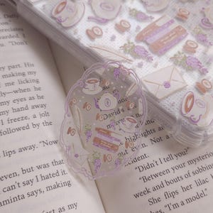 Könnte beinhalten: Ein transparenter Lesezeichen mit gewelltem Rand, verziert mit Abbildungen von Teetassen, Teekannen, Büchern und Trauben. Der Lesezeichen liegt auf einem offenen Buch, mit einer transparenten Handyhülle, die mit ähnlichen Illustrationen verziert ist.