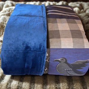 May include: A folded blanket featuring a solid royal blue plush side and a multi-patterned side. The patterned side includes sections of dark blue and grey plaid, a dark grey fabric with white lightning bolt icons, and a blue panel with a black raven.