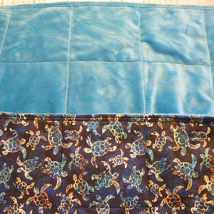 May include: A blue fleece blanket with a quilted design. The bottom edge of the blanket features a dark blue fabric with a pattern of colorful sea turtles.