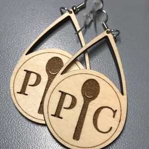 May include: A pair of wooden earrings with a teardrop shape. Each earring features a circle with the letters "P" and "C" and a stylized spoon design.