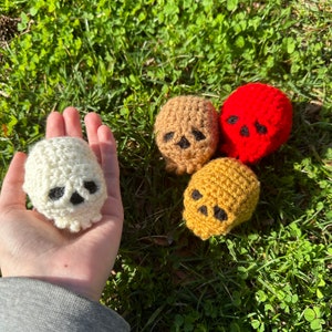 May include: Four crocheted skull ornaments in different colors: white, brown, yellow, and red. The skulls have black eyes and a black mouth.