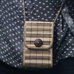 May include: A brown and beige woven crossbody bag with a brown button closure. The bag is hanging on a person wearing a black and white polka dot shirt.