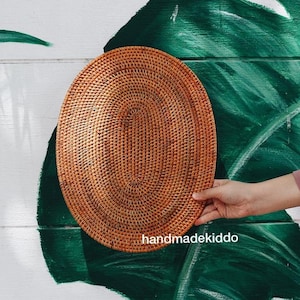 Könnte beinhalten: Ein geflochtener ovaler Teller aus natürlichem Rattan in hellbrauner Farbe. Der Teller wird von einer Hand vor einem grün-weißen Hintergrund gehalten.