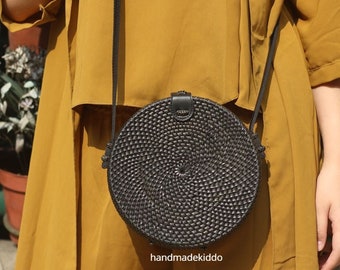 May include: A black round woven bag with a leather strap. The bag is worn over the shoulder of a person wearing a mustard yellow dress.