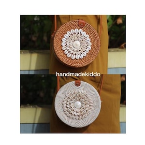 Peut inclure: Deux sacs ronds en paille tressée avec des accents de coquillages blancs. Les sacs ont des lanières en cuir brun et sont accrochés à l'épaule d'une personne.