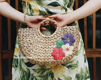 May include: A half-moon shaped woven straw bag with a round handle. The bag has colorful embroidered flowers on the front. The bag is handmade.