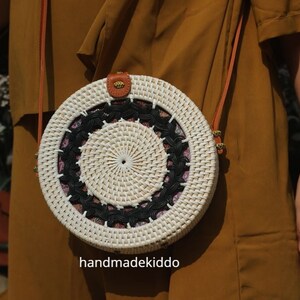 Puede incluir: Bolso bandolera redondo de rat&aacute;n blanco tejido, con un dise&ntilde;o circular en blanco y negro. El bolso tiene una correa de cuero marr&oacute;n y un peque&ntilde;o detalle de cuero en la parte superior. El texto "handmadekiddo" es visible.