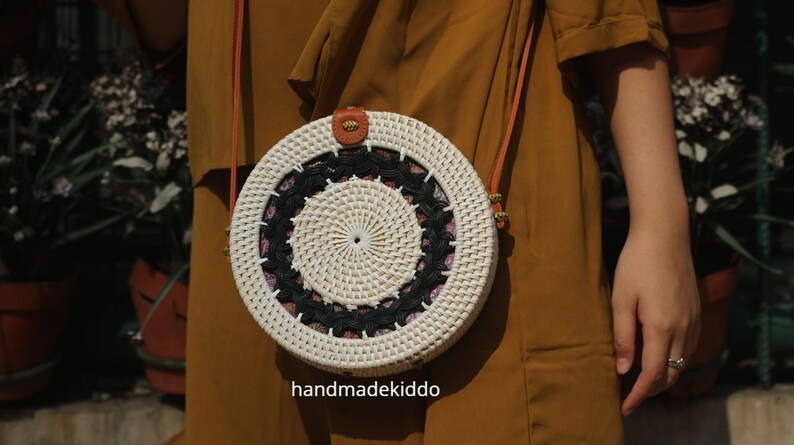 Puede incluir: Bolso bandolera redondo de rat&aacute;n blanco tejido, con un dise&ntilde;o circular en blanco y negro. El bolso tiene una correa de cuero marr&oacute;n y un peque&ntilde;o detalle de cuero en la parte superior. El texto "handmadekiddo" es visible.