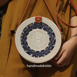 Puede incluir: Bolso redondo de rat&aacute;n tejido, con dise&ntilde;o blanco y azul marino. Cuenta con una correa y un cierre de cuero marr&oacute;n. El texto "handmadekiddo" es visible en la parte inferior del bolso.