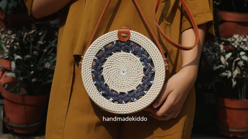 Puede incluir: Bolso redondo de rat&aacute;n tejido, con dise&ntilde;o blanco y azul marino. Cuenta con una correa y un cierre de cuero marr&oacute;n. El texto "handmadekiddo" es visible en la parte inferior del bolso.