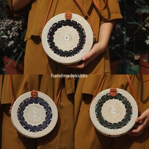 Puede incluir: Un bolso bandolera redondo de rat&aacute;n tejido con una correa de cuero. El bolso presenta un exterior blanco con un dise&ntilde;o central negro y estampado. El bolso lo sostiene una persona que lleva una blusa color mostaza. El texto "handmadekiddo" es visible.