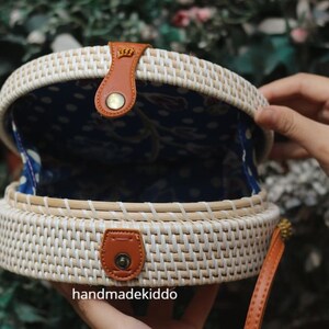 Puede incluir: Bolso redondo de rat&aacute;n tejido, blanco por fuera y con forro azul estampado. Tiene detalles de cuero marr&oacute;n, como la correa y el cierre. El texto "handmadekiddo" es visible en la parte inferior derecha.