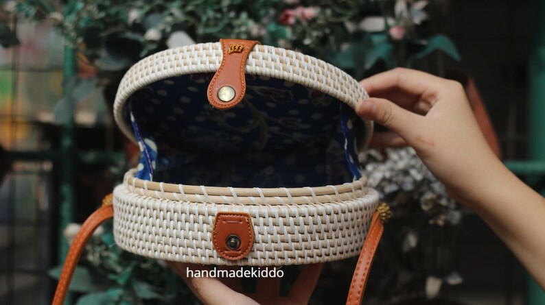 Puede incluir: Bolso redondo de rat&aacute;n tejido, blanco por fuera y con forro azul estampado. Tiene detalles de cuero marr&oacute;n, como la correa y el cierre. El texto "handmadekiddo" es visible en la parte inferior derecha.