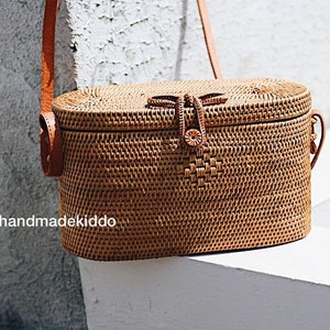 May include: A brown woven oval-shaped handbag with a leather strap and a woven closure. The bag is sitting on a white surface. handmadekiddo